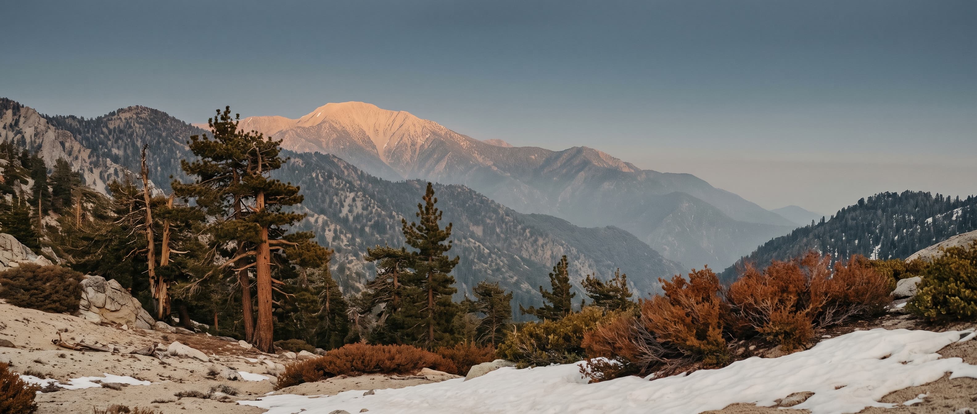 Alpenglow on a high San Bernardino ridgeline at golden hour, layered pine-covered ridges receding into alpine haze with Jeffrey pines, granite, and patches of snow — the same alpine stillness that connects Big Bear to Nagano